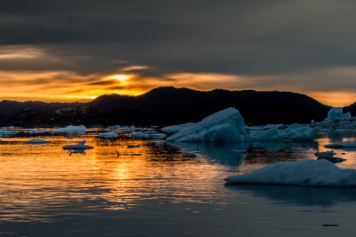 De ondergaande zon kleurt de fjord en de drijvende ijsbergen oranje.