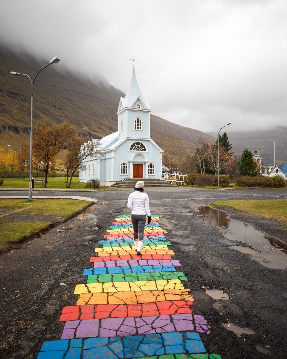 Een wandelaar loopt over een regenboog pad richting het kerkje van Seydisfjördur.