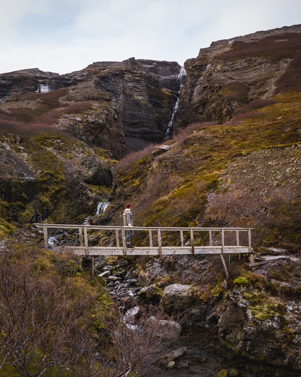 Een vrouw kijkt vanaf een houtenbrug en wandelpad uit op de Valagil waterval in de Westfjorden van IJsland. 