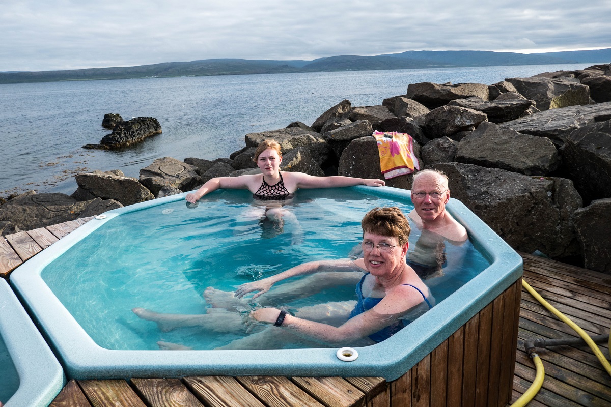 Reizigers genieten van de hottub in Drangsnes in de Westfjorden, gelegen in IJsland. Het badje ligt direct aan zee wat een fantastisch uitzicht geeft. De zon schijnt.