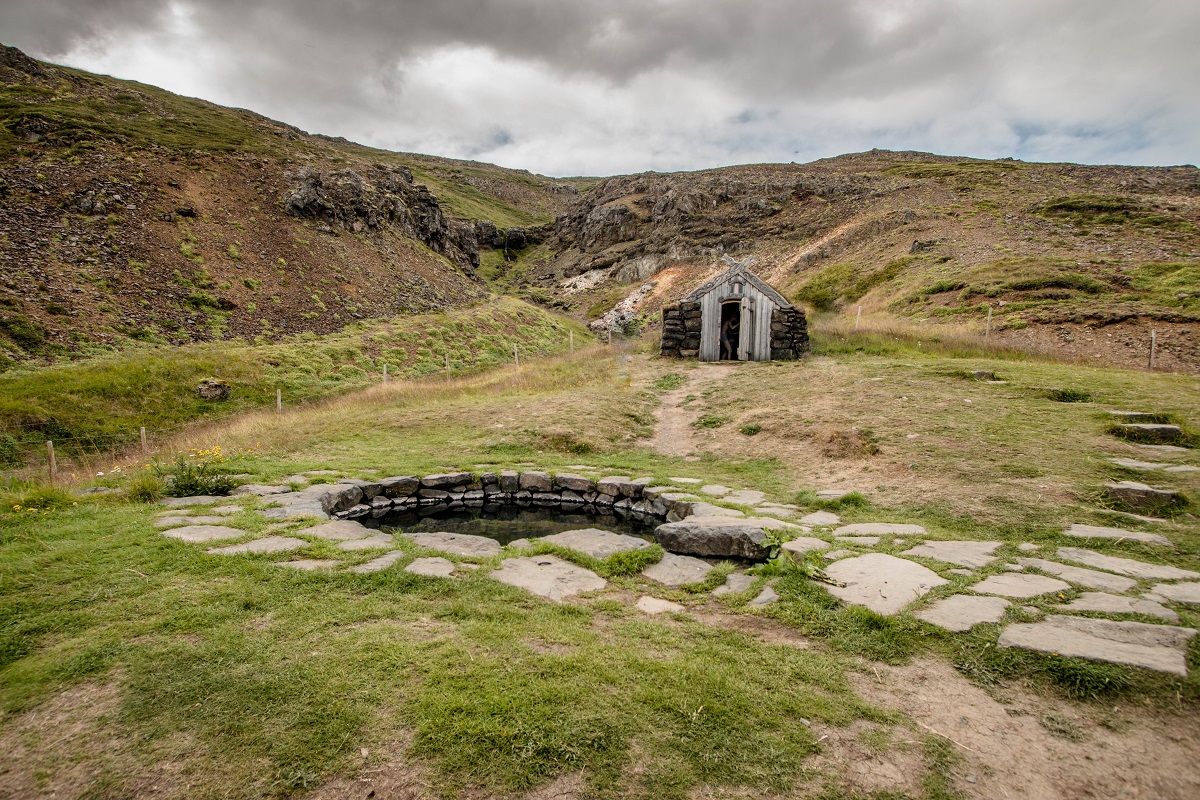 Het geothermische badje Gudrunarlaug in het met mos bedekte landschap. Op de achtergrond staat een reiziger zich om te kleden in het houten hutje.