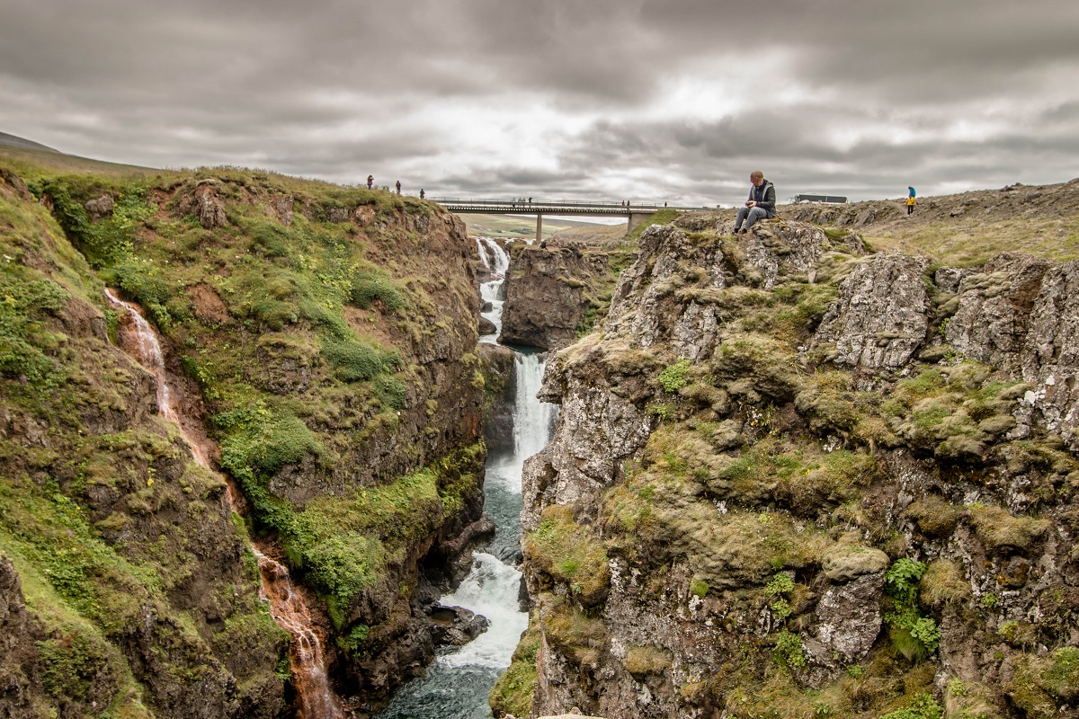 Een waterval loopt door de Kolugljufur kloof in het noorden van IJsland. Reizigers zitten op de rotsen aan de rand van de kloof en staan op de hangbrug.