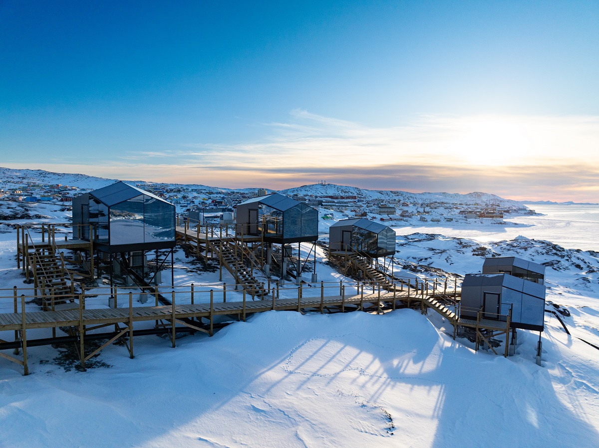 De hutten van Aurora Cabins staan in een besneeuwd winters landschap. De reflectie op de hutten laat de uitzicht op het fjord zien. Vanaf de boardwalk loop je naar de cabins toe. Op de achtergrond zie je Ilulissat.