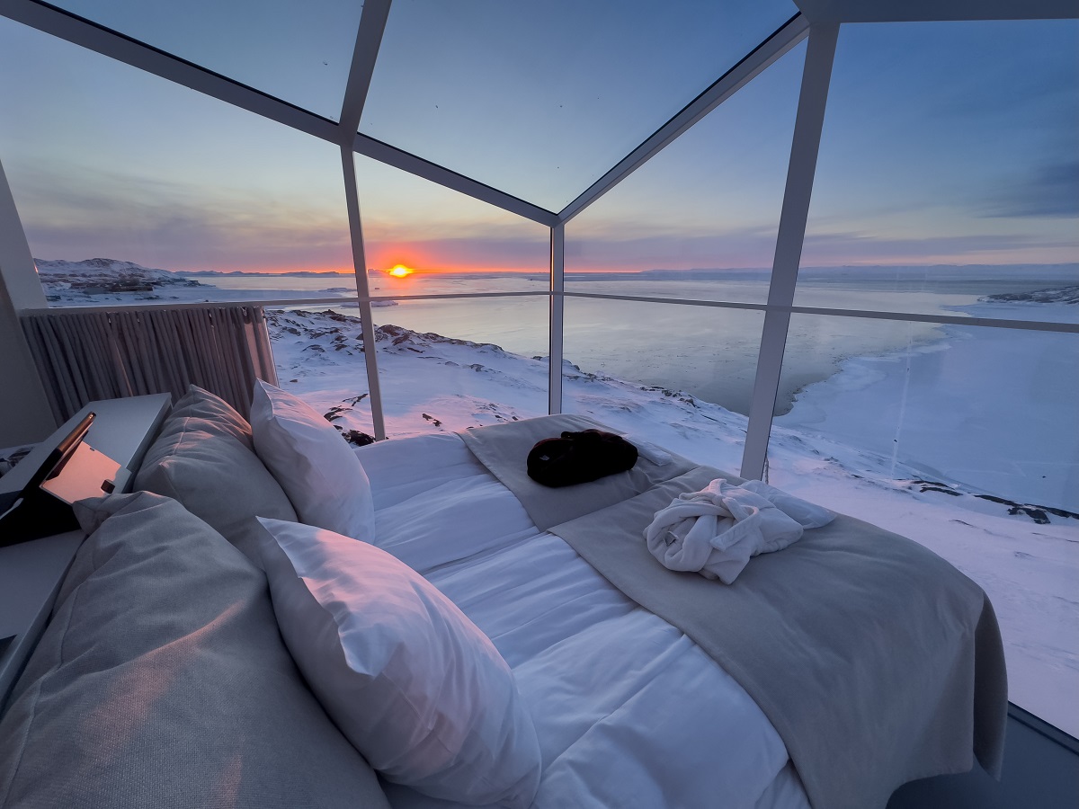 Het uitzicht vanaf het bed in een van de Aurora Cabins over het winterse Groenlandse landschap. Vanuit de kamer kijk je uit op de fjord en zonsondergang.